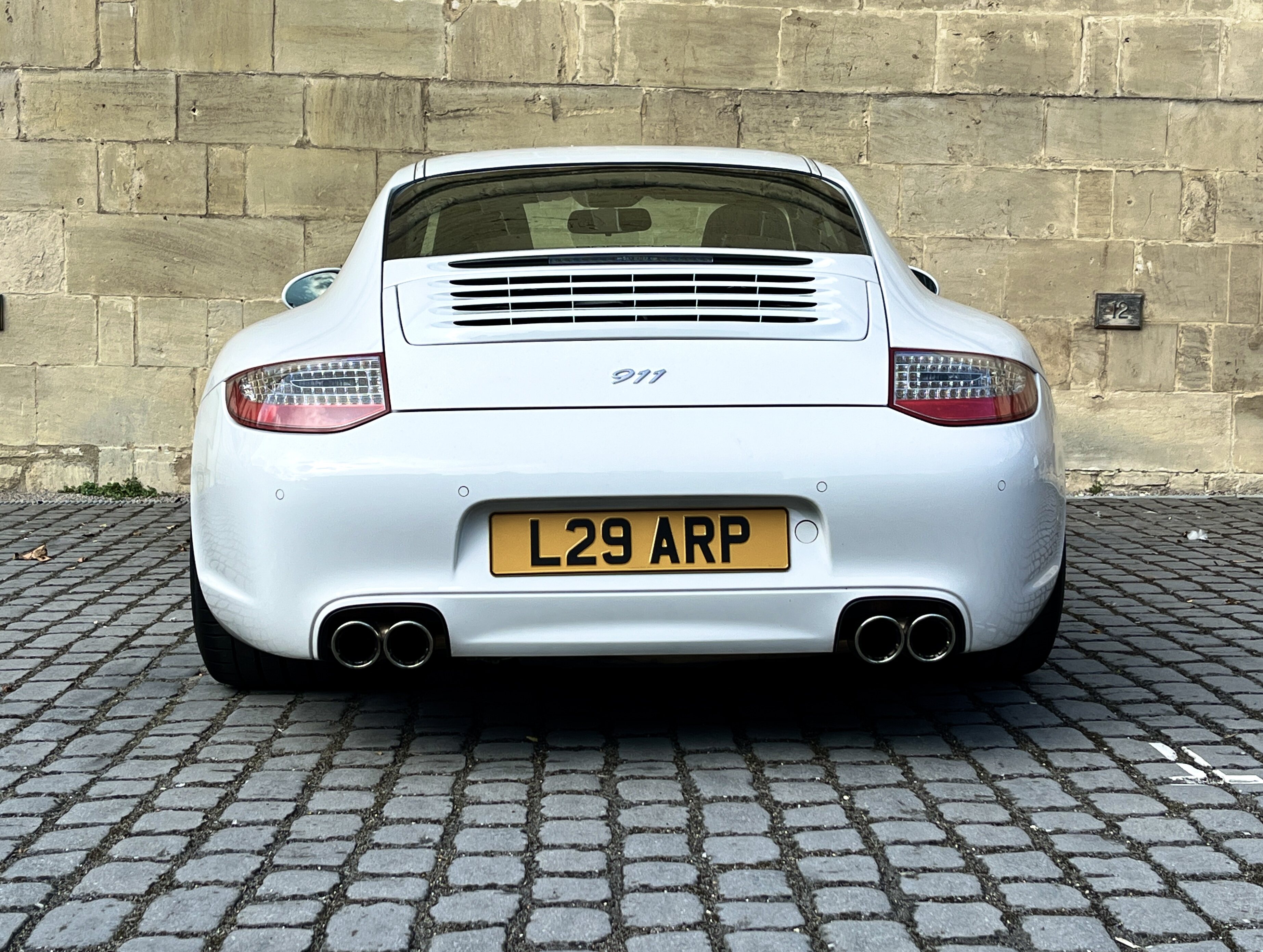 Pistonheads - The image depicts a white sports car parked on the side of a street. The car is positioned at an angle, showcasing its sleek design and the license plate that reads "L26 ARP." There's a clear view of the rear side of the vehicle where the exhaust pipes are visible. The setting appears to be urban, with cobblestone pavement and a building in the background. The overall image gives off a sense of luxury and high-performance vehicle.