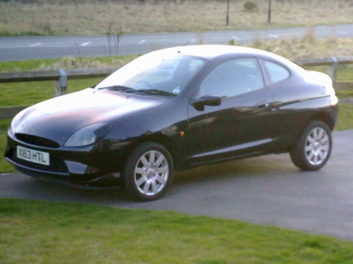 My Car - Page 1 - Readers' Cars - PistonHeads - The image shows a sleek, dark-colored coupe parked on the side of a street. The reflective angle of the photograph enhances the shine on the car's surface. The vehicle is parked near the curb, and a lighting fixture can be seen on a nearby structure. The street appears to be peaceful, with no visible traffic or pedestrians. The car is positioned in such a way that part of its rear is visible, revealing its distinctive design and style.