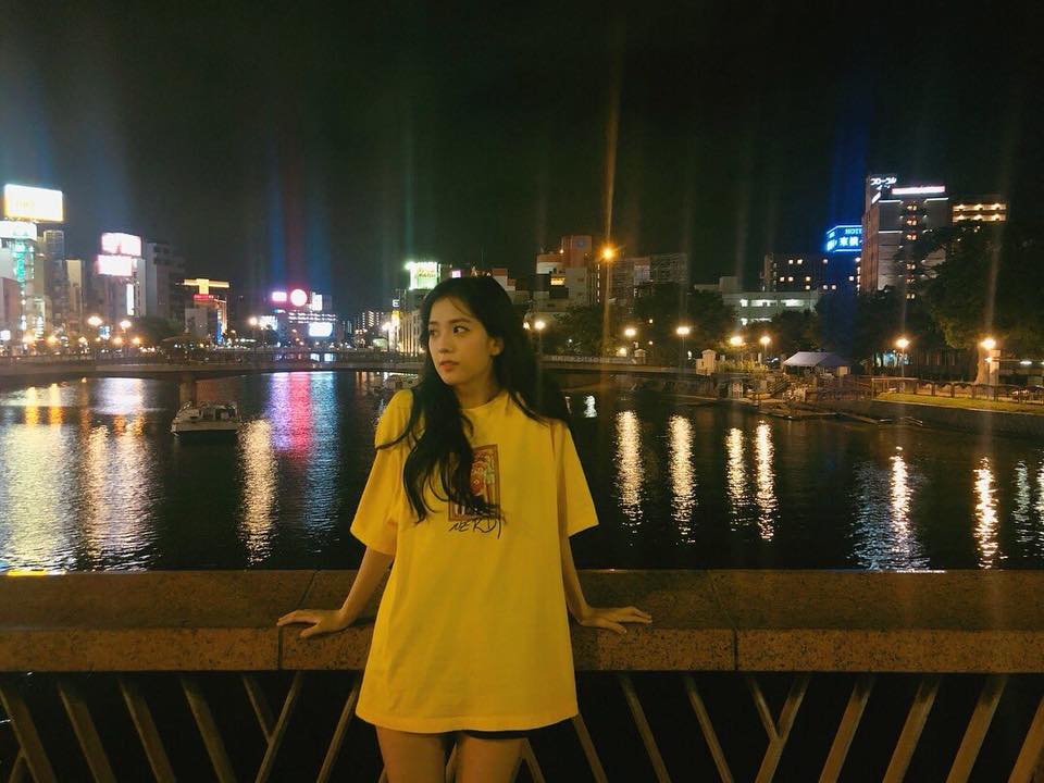 A woman standing on a pier with her arms crossed - The image features a young woman standing on a bridge at night with a lit-up cityscape in the background. She is wearing a yellow blouse and appears to be looking back at the viewer. The bridge overlooks calm water, possibly a river or part of a riverway. In the background, vibrant lights from buildings and streetlamp illuminate the surrounding area, creating a picturesque urban night scene.