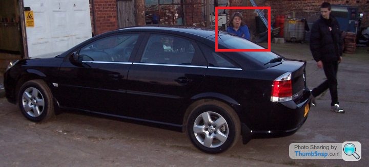 Pistonheads - The image shows an outdoor scene, likely a private property based on the presence of a white door and brick walls. There is a man walking past the car, notably dressed in a black hoodie. Parked on the left side of the image is a grey-colored hatchback vehicle. Across the back windshield, a man appears to be driving the car. The car's back section is visible and noticeably pulled out, possibly to load or unload cargo. The woman in the image is not interacting with the car or the man in the black hoodie, but rather seems to be waiting or watching.