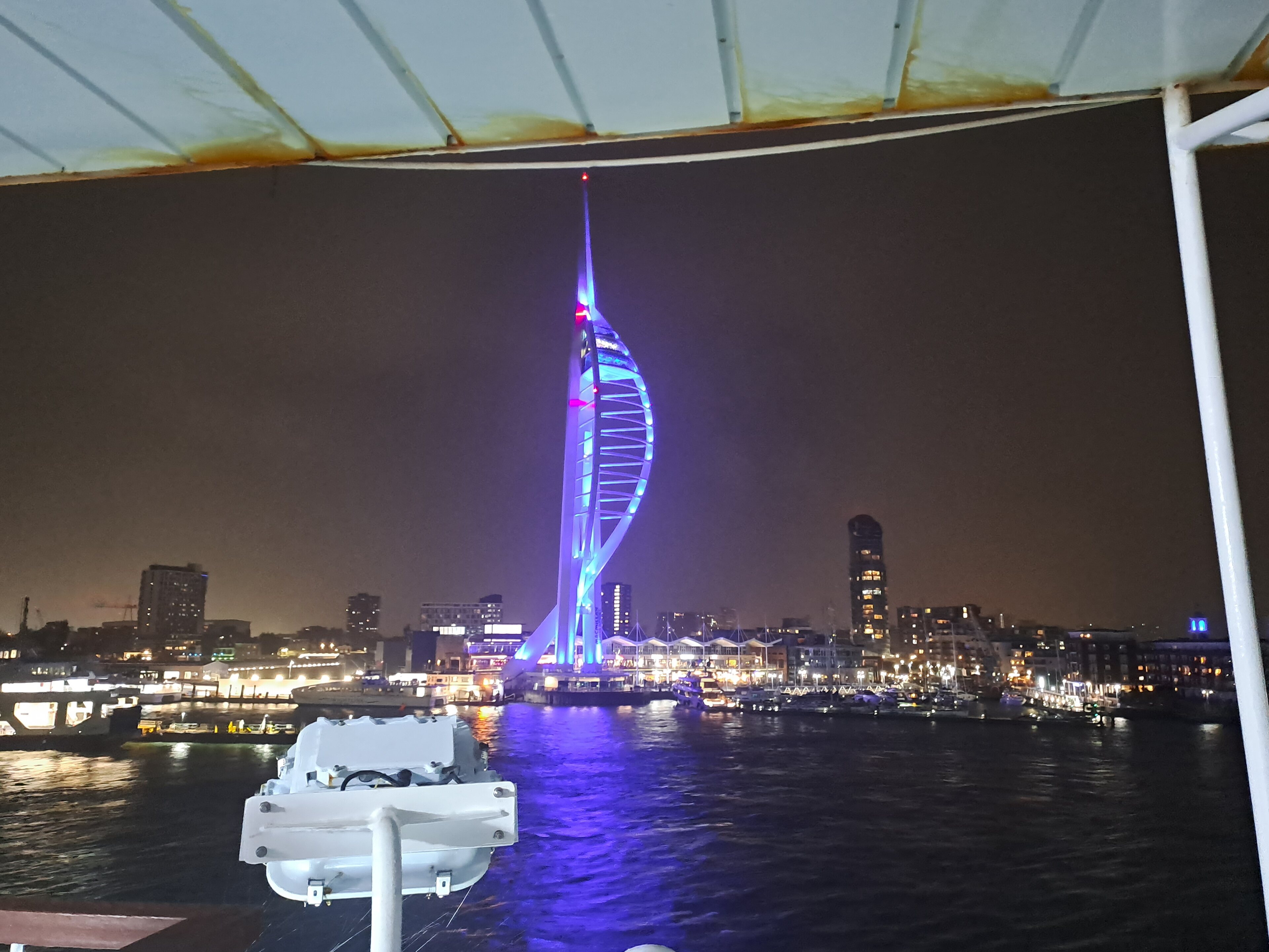 Pistonheads - The image captures a nighttime view of a cityscape, with a vibrant blue light emanating from a building in the distance. Prominently featured in the background is a large Ferris wheel, suggesting that the location might be a tourist destination or near an amusement park. In the foreground, a boat is docked, its silhouette visible against the city lights. The sky above is dark and devoid of stars, indicating a cloudy night.