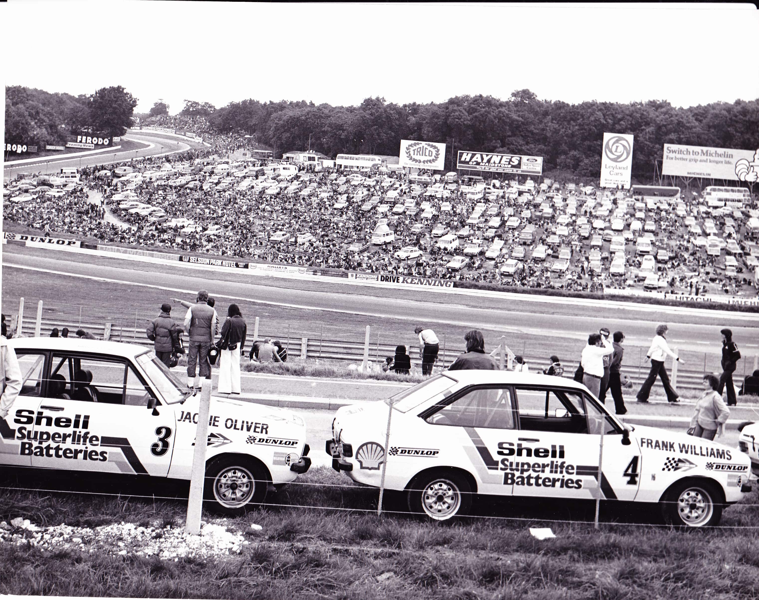 How about a 'period' classics pictures thread - Page 500 - Classic Cars and Yesterday's Heroes - PistonHeads - This image appears to be a black-and-white photograph capturing a moment from a public event or sports competition. It shows a large crowd of spectators filling the stands. In the foreground, several vintage cars, which have Shell and Super Life Batteries sponsorship logos, are parked alongside each other, suggesting that they might be on display or associated with the event taking place. A few people can be seen walking between or near the cars, indicating a casual, relaxed atmosphere at the venue. The setting seems to be a racetrack or outdoor event space, as evidenced by the fencing and open sky visible in the image.