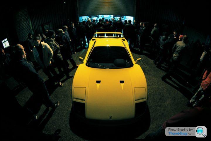 The GT Cafe - ace cafe of the north - FEB Meet - Page 1 - North West - PistonHeads - The image depicts a dimly lit room giving the vibe of a clandestine setting. A crowd of people is gathered around a large sports car that is prominently displayed in the center of the room. The car appears to be a Lamborghini, characterized by its sleek design and pointed front end. The atmosphere suggests an event or gathering, possibly an exclusive car show or viewing. The use of warm, ambient lighting casts shadows and highlights, emphasizing the car's features and adding to the overall mood of the scene.