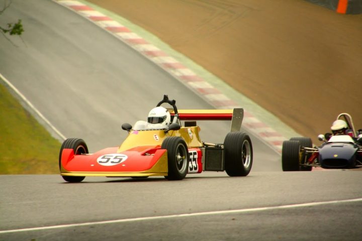 A motorcycle is parked on the side of the road - Pistonheads - The image displays an exciting scene from a racing track. At the center of the action is a small gold race car, likely a single-seater, driven by a person donning a white helmet. The car is moving at high speed, leaving a clear gap between it and the second car in the background. This car has a more modern design with a prominent front wing, and its driver is also wearing a white helmet. The racing track is a standard circuit with curbs and barriers visible in the background. The overall atmosphere is one of intense competition and high-speed racing.