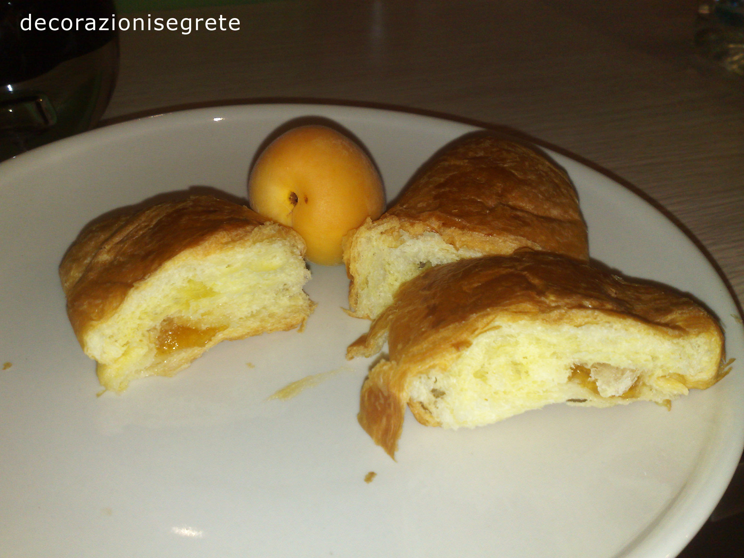 The image shows a white plate with three pieces of baked bread. They appear to be partially eaten, revealing the interior's flaky texture. Situated among the bread is a whole peach, resting comfortably on the plate. The text "decorazioneisegregrete" is written in the upper left corner, but without context, its purpose in this image remains unclear. The overall scene suggests a simple yet pleasant meal.