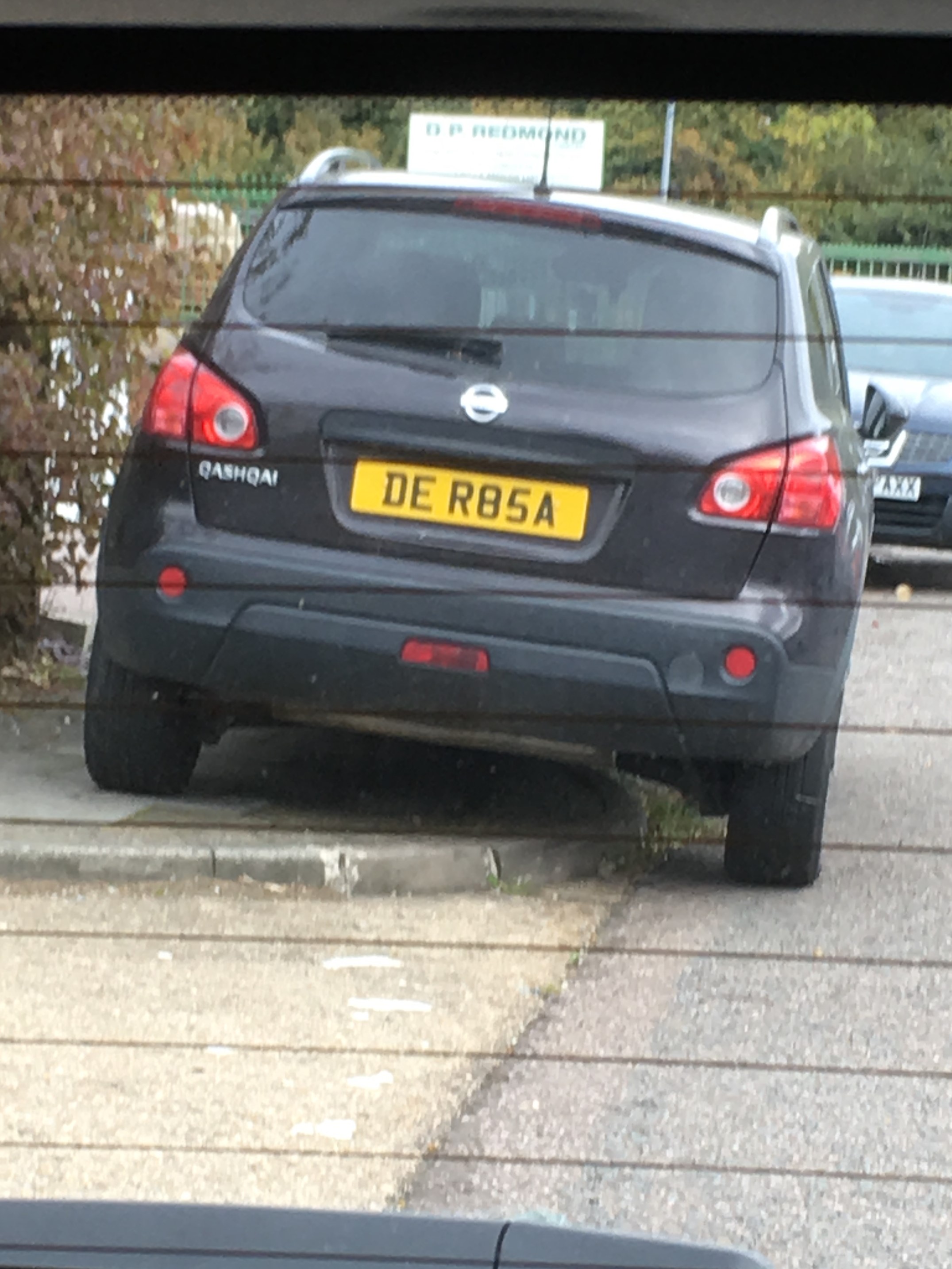 What C124PPY personalised plates have you seen recently? - Page 264 - General Gassing - PistonHeads - This image depicts a rear view of a parked car captured through a grid or metal mesh, possibly the window of another vehicle. The car is dark-colored with a visible European-style license plate that reads "DE R85A." The vehicle has tinted windows, and there is visible sunlight indicating it may be daytime. In the background, there are hints of a garden and additional vehicles, but they are less distinct from the parked car itself.