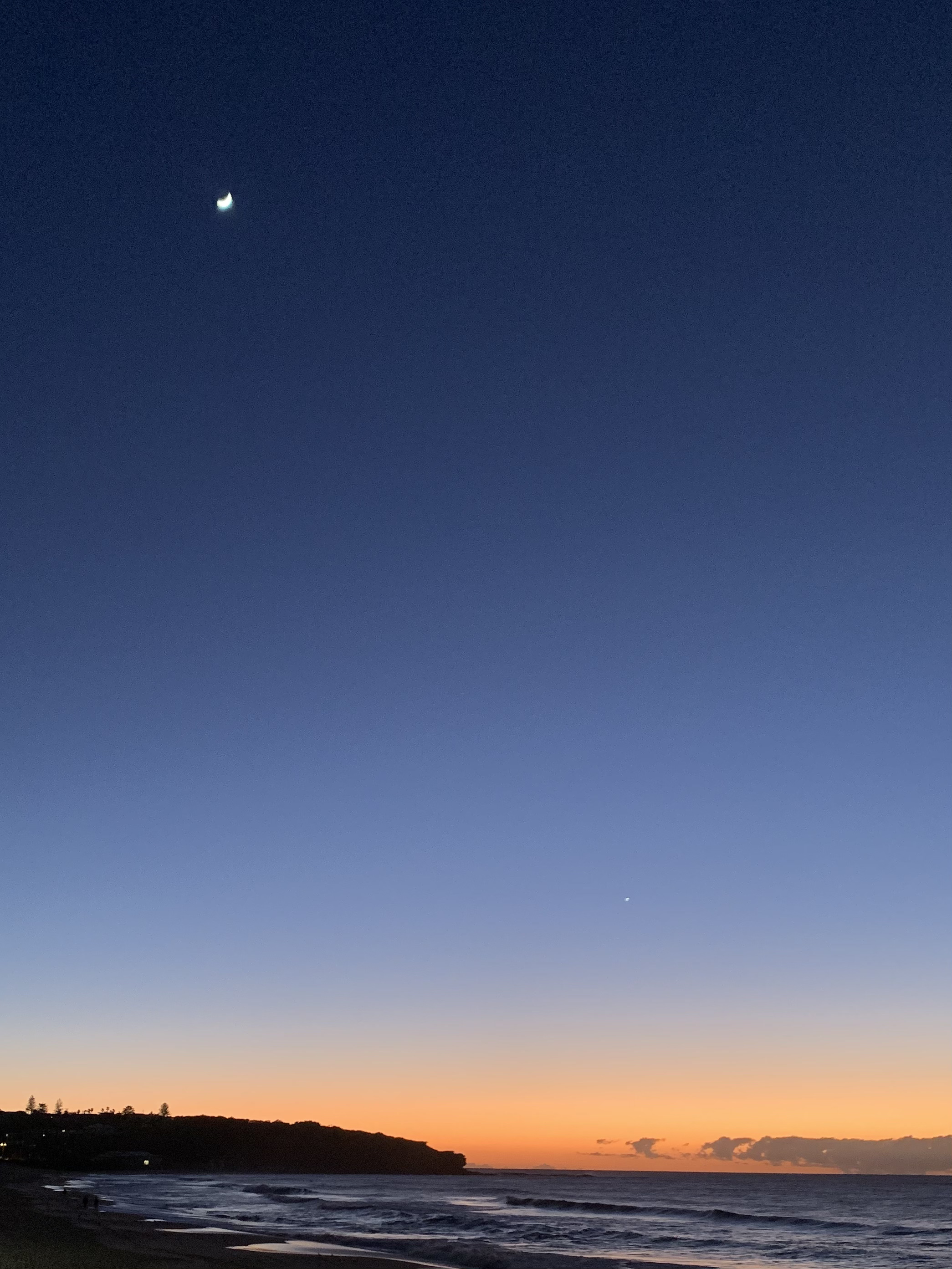 April 2021 Photo Competition - Above the Horizon - Page 1 - Photography & Video - PistonHeads UK - The image captures a serene night scene at a beach. A crescent moon is visible in the clear sky above, adding a touch of celestial beauty to the scene. Below the moon, the horizon is illuminated by the warm glow of the city lights and streetlights, which seem to be reflected on the water's surface. The sandy beach stretches out towards the horizon where the cityscape meets the ocean. There are no visible people or objects, adding to the tranquil atmosphere.