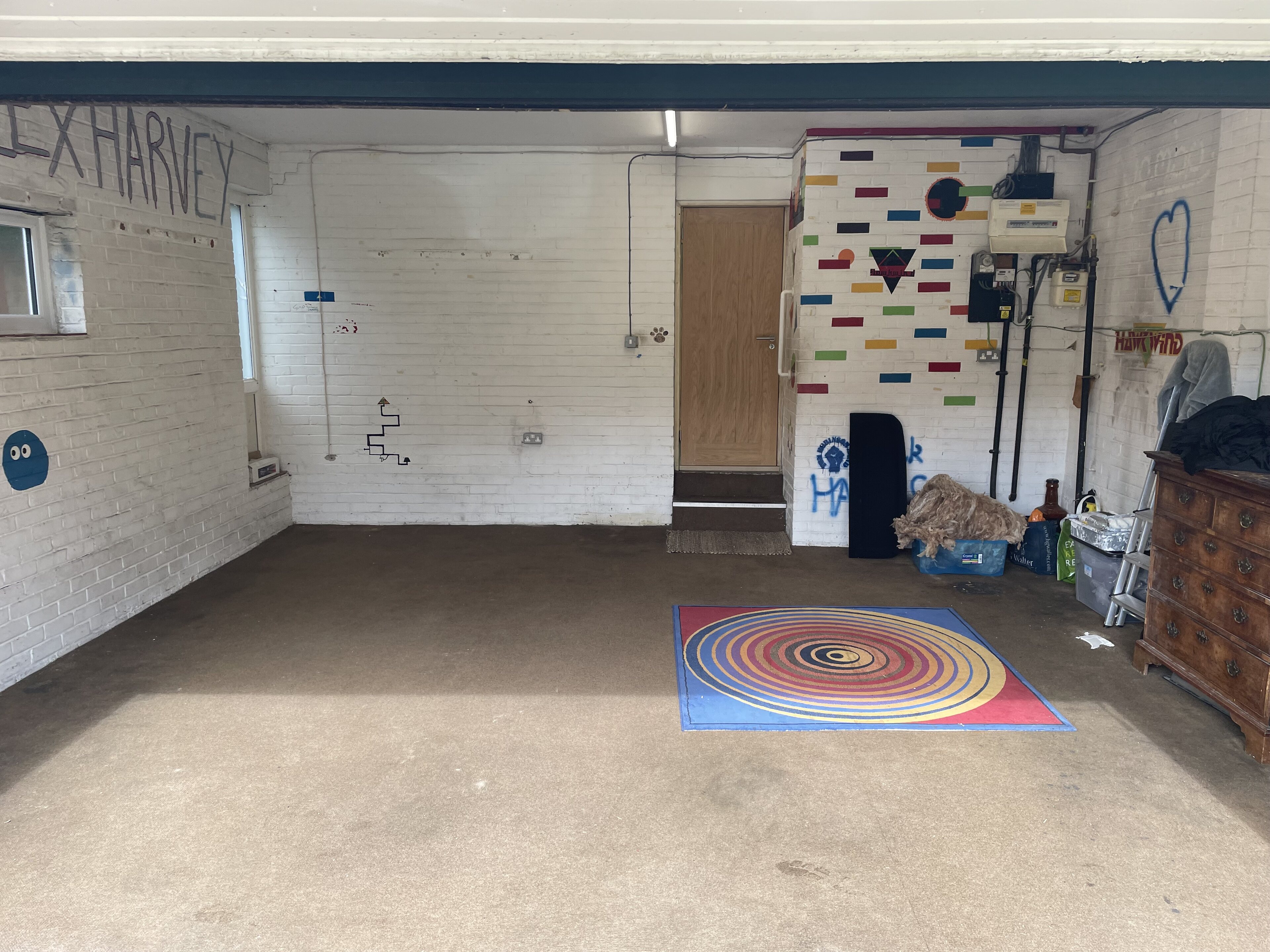 My garage improvement  - Page 1 - Homes, Gardens and DIY - PistonHeads UK - This is an image of a garage with a large open door. The interior of the garage is quite spacious, with concrete flooring and white walls. On one side, there's a black and white rug or carpet laid out on the floor, featuring a colorful swirl design. To the left, there's a wall with a sign that reads "Harris," indicating the name of the owner or the property. The garage is equipped with various pieces of machinery or equipment, including what appears to be a large piece of metal, possibly part of industrial or mechanical equipment. On the right side of the image, there's a small object that looks like a heart-shaped item hanging on the wall. The overall appearance suggests that this garage might be used for various purposes such as storage and perhaps work on mechanical or construction projects.