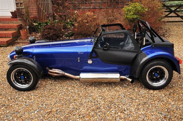 R500 K Series - Page 1 - Caterham - PistonHeads - The image shows a small, convertible-top sports car parked on gravel. It is painted in a unique shade that combines blue and black hues. The car has one door visible, and its roof appears to be down. There are large, chrome exhaust pipes extending from the back of the vehicle, suggesting it might have a powerful engine. The vehicle's setting includes a paved driveway, planted greenery, and some bricks, which hint at a residential area.