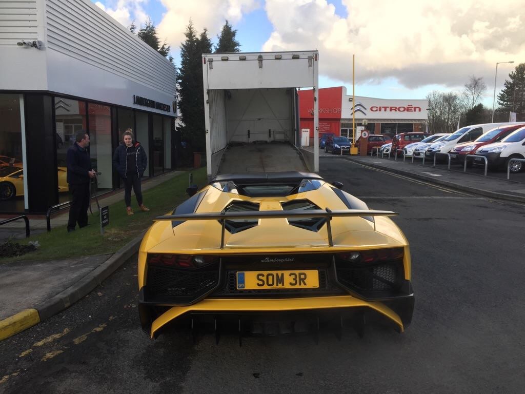 Aventador SV Register - Page 2 - Supercar General - PistonHeads - The image captures a moment in an outdoor parking lot where a striking yellow supercar is parked. The car features a long hood and short rear, with two large air intakes at the back. The license plate reads "SOM M3R". To the driver's side, several pedestrians are standing by, possibly spectators or employees of a nearby building. The car is accompanied by a white delivery truck with the door open and no visible cargo. In the background, there is a partially obscured building with a red sign, and beyond it, other vehicles are parked, creating a lively contrast with the sleek and exclusive supercar.