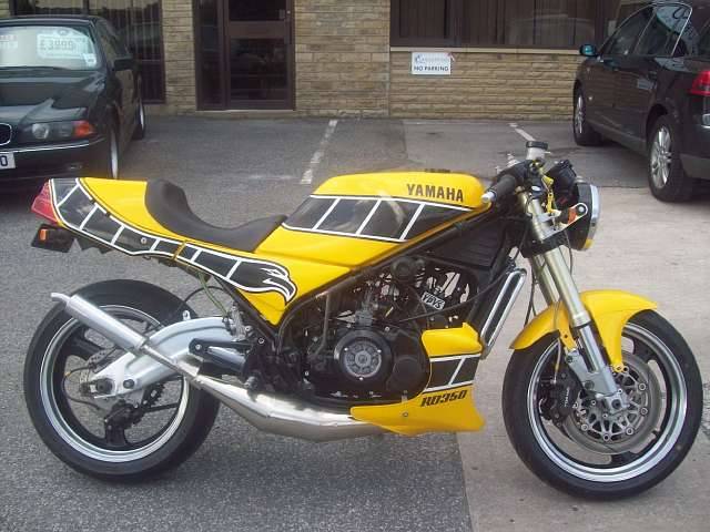 Pistonheads - The image showcases a uniquely decorated yellow Yamaha motorcycle parked on an open asphalt surface, possibly in a parking lot. The motorcycle's bodywork features large pieces of black and white design, similar to a cartoon-style eagle motif, which is prominently displayed on both the front and rear panels. Aside from the motorcycle, there are a few other vehicles, including a black car and a few more in the background that are partially obscured. The setting suggests a public area, and the focus is clearly on the motorcycle with its distinctive design.