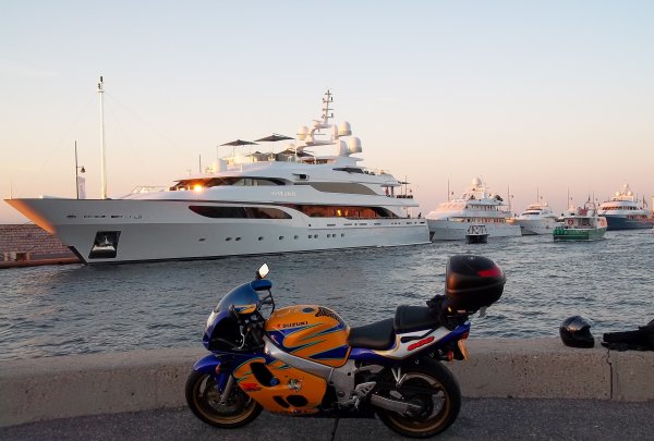 Universal panniers - Page 1 - Biker Banter - PistonHeads - The image captures a serene marina scene during dusk. A large white luxury yacht is docked, its size and presence suggesting a sense of grandeur and luxury. Several smaller boats are also seen in the water, some closer to the yacht and others further away. On the pier, a blue motorcycle is parked, reflecting the light and adding a touch of modernity to the scene. The overall atmosphere is calm and peaceful, with the yellow glow of the sunset casting over the scene, creating gentle shadows and highlighting the tranquility of the water.