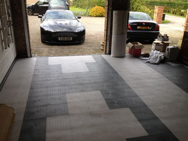 Garage Queens - Page 9 - Aston Martin - PistonHeads - The image depicts an indoor residential space that leads out to a gravel driveway. There are two cars parked in the driveway - a black sports car on the left and a dark-colored sedan on the right. The carpets of the room feature a diamond-square-and-half pattern, running parallel to the door opening. The driveway, easily visible through the doorway into the garage or house, features an interlocking brick pattern, and the sky is visible outside the garage door, suggesting it may be an early morning or late afternoon scene.