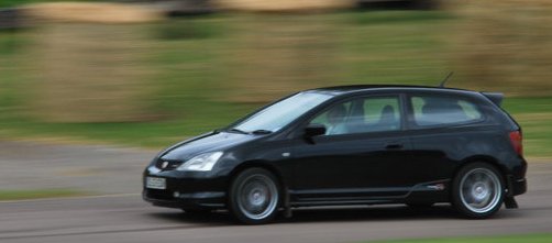 Type R - Page 1 - Honda - PistonHeads - This is a dynamic image capturing a small black car speeding down a curvy paved road in a blurred, almost motion-picture style effect. The car is seen from the rear, passing by a grassy area with hay bales. The background is a slightly blurred, out-of-focus greenery, giving the impression of speed. There is no visible text or distinctive branding on the car that can be discerned from this angle. The image conveys a sense of movement and energy.