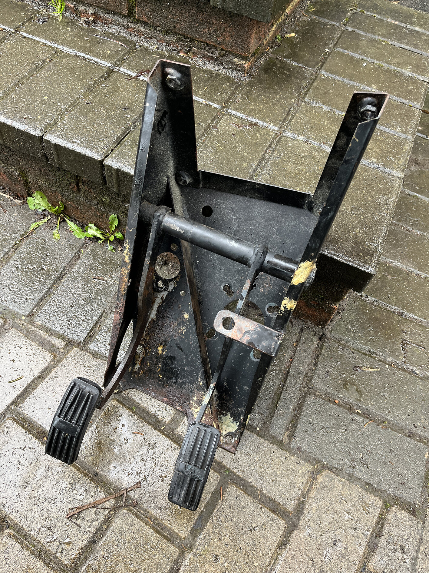 Pistonheads - The image shows an old, rusted metal object that appears to be a clamp or attachment device. It is located outdoors on what seems to be a wet, possibly rain-soaked surface. The object has several parts: a main body with a wheel and two handles, suggesting it might have been used for lifting or moving heavy items, like construction materials or machinery. There are also metal brackets attached to the main body, which may have been part of a larger system that is now disassembled or removed. The rust indicates that this object has been exposed to the elements for an extended period and has likely not been used in many years. The surrounding environment is quite nondescript, with no distinctive features or landmarks that provide context or location.