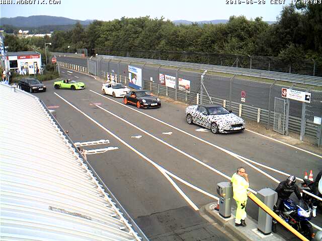 Ring Pistonheads Webcam - The image depicts a bustling scene at an airport or large parking facility. Various modes of transportation are visible, including cars, a motorcycle, and bicycles. People are scattered throughout the scene, walking or standing, possibly waiting or just passing through. The ground is a mix of tarmac and concrete, and a fence separates the paved area from a lush green hillside in the background. Buildings can be seen in the distance, further suggesting the proximity to an airport or transportation hub. The image appears to be taken with a timestamp at the top-right corner, indicating it might be a screenshot from a security camera or a traffic monitoring system.