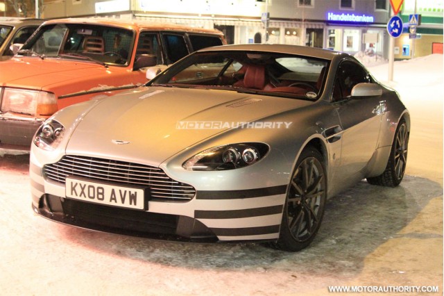 Vantage Pistonheads Side Bumpers Skirts - The image showcases a striking silver Aston Martin sports car on a snowy street during a business trip. The car, adorned with vertical stripes, stands out against the white backdrop. A few pedestrians are visible in the background, further emphasizing the urban setting. The car's sleek design and polished surfaces are even more pronounced on the glossy surface of the snow.