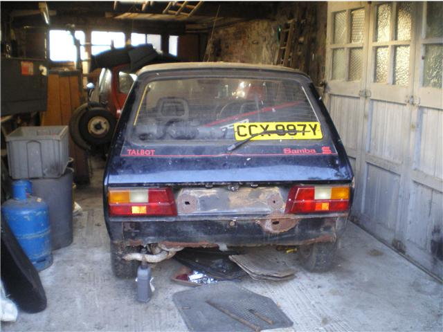 Anyone remember the Talbot Samba S? - Page 1 - Classic Cars and Yesterday's Heroes - PistonHeads - The image shows a blue car parked in a garage. The car appears to be an older model, possibly a compact car or a minicab, as indicated by the size and the label "Samba 500" on the trunk. The taillights are intact, with the license plate beginning "CXX 097Y." The garage is dimly lit, and in the foreground, you can see a variety of objects scattered around, including what looks to be a metal bucket and a large blue gas cylinder. The presence of mechanical or industrial equipment in the background suggests the garage may be used for repairs or modifications.