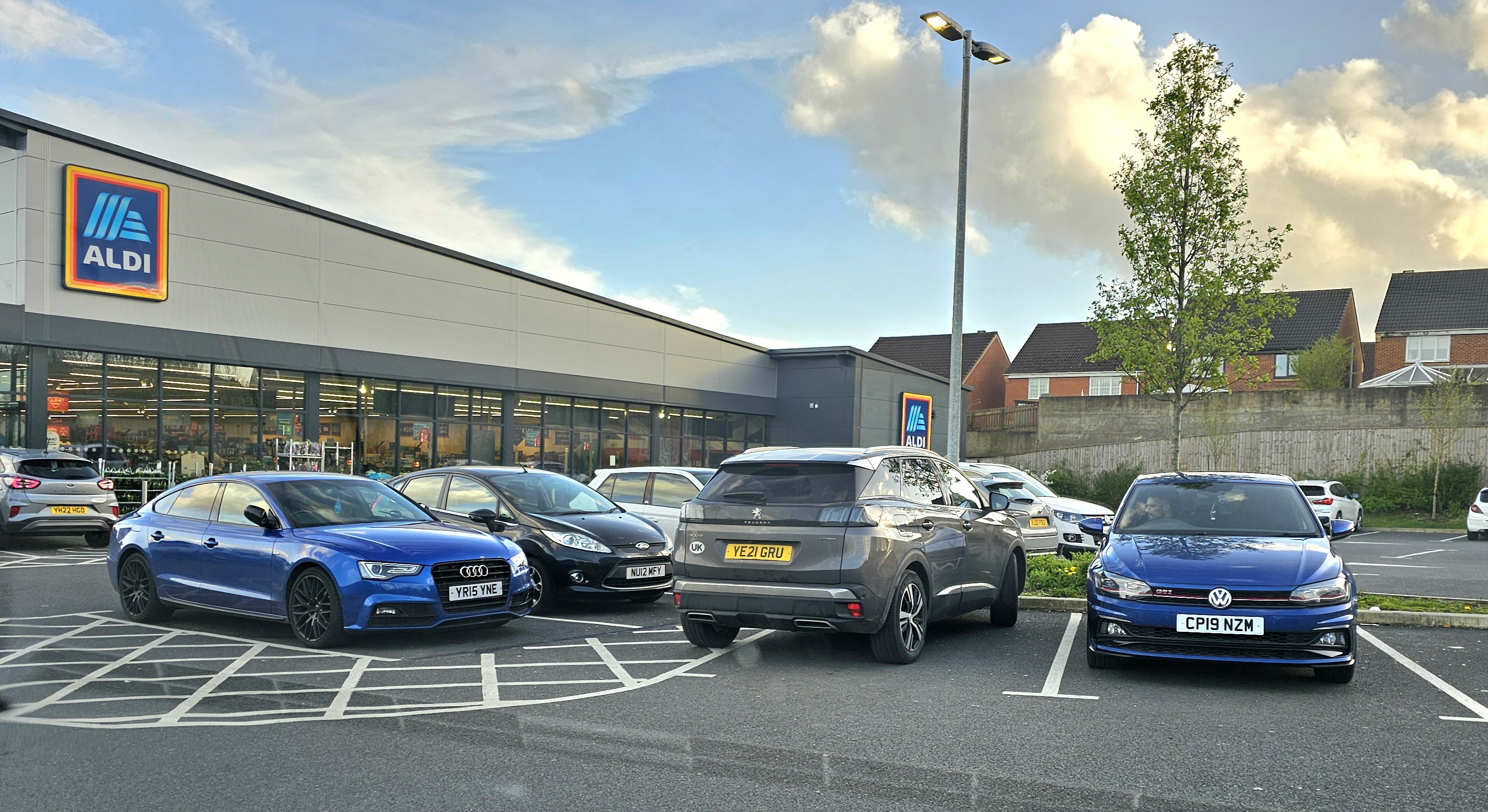 Pistonheads - The image shows an outdoor scene with a large car dealership in the background. In the foreground, there are several cars parked by the roadside. The sky is cloudy and the time of day appears to be late afternoon or early evening. There is a sign indicating the presence of a car dealership, and the lot is well-maintained with clear markings for parking spots.