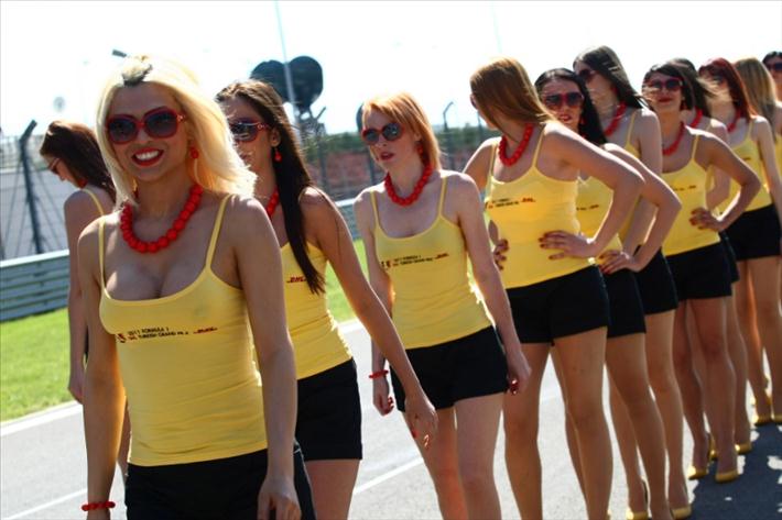 Grid Babes Girlspit Pistonheads - The image shows a group of women standing in a line on a sidewalk, all dressed in matching bright yellow tank tops, black short shorts, and red necklaces. They are all smiling and appear to be ready to perform, as suggested by the dance attire. The environment suggests they may be at a race track, given the grassy background and the placement of the women along what looks like a race car track. Each woman is wearing matching red sunglasses, giving the group a coordinated and vibrant look.