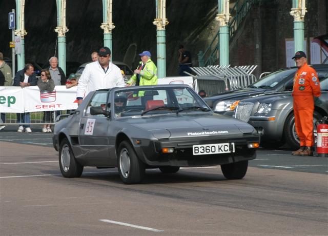 Beautiful Bertones Pistonheads - The image depicts an outdoor scene where a man is walking past a grey sports car with a convertible top down. He is dressed in an orange racing suit, complete with a helmet, and is standing near a large fire extinguisher. This man appears to be in a race car driver's outfit, indicating he might be a driver or someone involved in motorsports. The sports car is positioned in the middle of the image, capturing the viewer's attention. The surrounding cars and the presence of a crowd suggest this might be a public event, possibly a car show or a racing event. There is a sense of action and anticipation, given the man's attire and his proximity to the sports car.