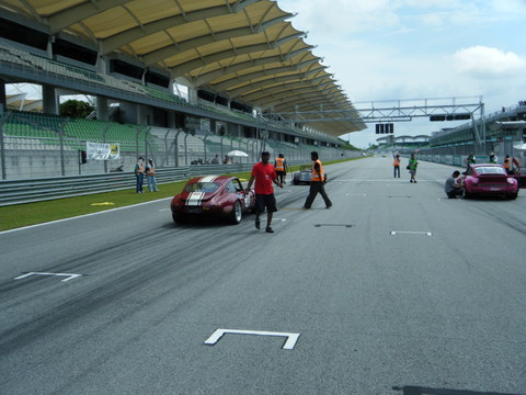 Pistonheads Afos Sepang - The image depicts a scene at a Formula 1 racetrack. There are several people present in the stands and at the trackside, some possibly racers or team members. A red car is driving on the track, perhaps a part of a test run or a practice session. The arena has a curved ceiling with yellow supports, possibly to protect spectators from debris or to shield them from the elements. The atmosphere is focused and professional, indicative of a pre-race activity or a training exercise.