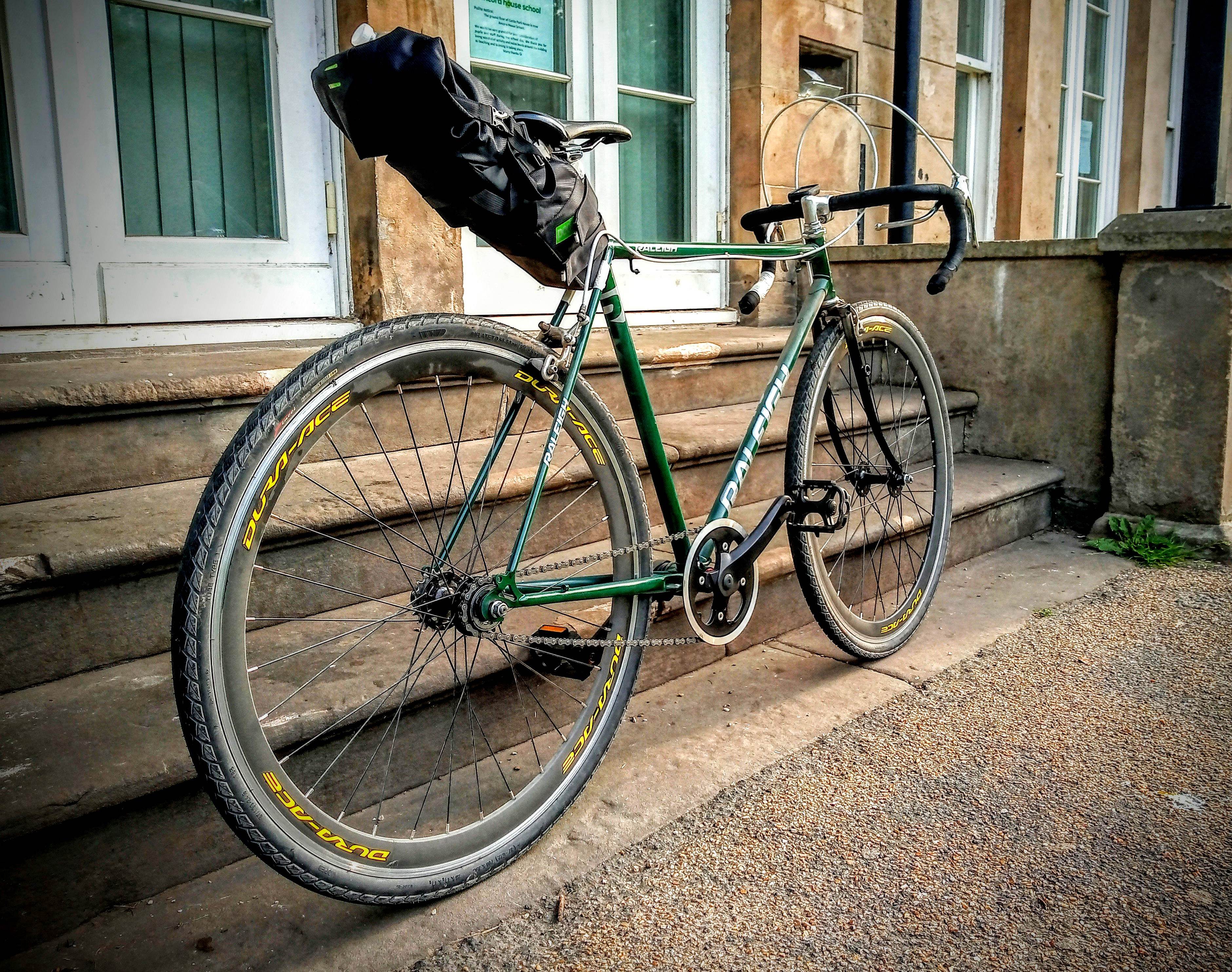 Retro Road Bikes ? - Page 8 - Pedal Powered - PistonHeads UK - The image showcases a vintage bicycle parked against a brick wall. The bicycle has a striking red frame and features fenders, which is indicative of older models. It's equipped with black tires and yellow wheels. The handlebars are adorned with various accessories, including a bell and a pair of water bottles, suggesting that this bike is used for long rides or touring. A helmet is casually hung on the handlebars, implying recent use. The bicycle takes up most of the frame from left to right, giving it prominence in the image.