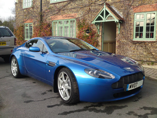 Arggh Clutch - Let the mods begin! - Page 1 - Aston Martin - PistonHeads - The image shows a blue Aston Martin car, which appears to be a modern and upscale model. The car is parked on a road next to a brick building with green shutters. The car has a sleek design with a curved front and large, shiny wheels. There is a license plate attached to the front bumper of the car. The setting suggests a residential area, possibly in an urban neighborhood with well-maintained architecture. The environment looks orderly and clean, suggesting a well-kept neighborhood.