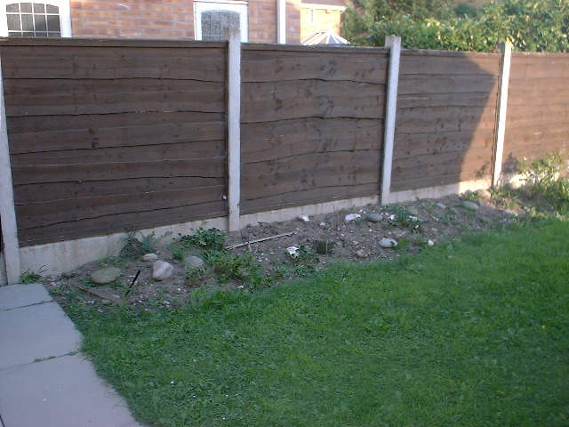 A bench in front of a brick building - Pistonheads - The image shows a wooden fence, which appears to be in a garden or outdoor area. The fence is composed of vertical boards, each showing signs of wear. The front of the fence, facing the camera, shows a somewhat worn appearance, with the paint or stain chipping away or being worn off. There are rocks scattered around the base of the fence. The background reveals a glimpse of a building and more fencing, but the focus of the image is the garden area with the wooden fence. There are no visible people or animals in the area, and the lighting suggests it might be late afternoon or early evening.