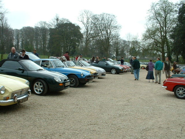 FAO MGJohn - Page 1 - MG - PistonHeads - The image captures a scene at a vintage car event. The ground is covered in gravel, and several vintage cars are parked neatly in rows. Each car is distinct, representing various eras and styles. The people present are spread throughout the area, some standing closer to the cars, while others are further away. The trees in the background provide a natural border to this outdoor event. The sky above is overcast, casting a uniform light over the scene.