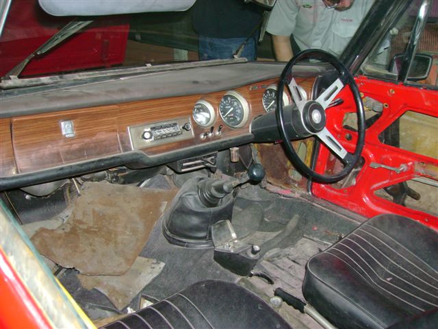 Beautiful Pistonheads Bertones - The image shows an interior view of a police car's cabin. The car features a vibrant red exterior and black leather upholstery. The dashboard is a symmetrical wood panel with chrome accents, housing multiple gauges and a clock. A large steering wheel and gear stick are central on the dashboard. The interior seems weathered and well-used, indicating frequent use. The ambient lighting inside the car suggests a setting that is neither too bright nor too dim.