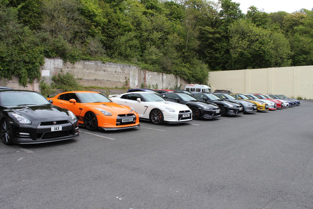 Pistonheads - The image captures a serene parking lot scene set against a concrete wall covered in lush greenery. The lot is filled with ten cars, all parked neatly in a row. The colors of the cars vary from black, white, and orange, adding a vibrant touch to the otherwise monotonous backdrop. A medical van stands out amidst the cars, its distinct white and blue colors marking it from the rest.