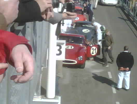 Goodwood Revival Who is going - Page 1 - Classics - PistonHeads - The image captures an exciting scene from an auto race. The focus is on a vintage red race car with the number 3 on it, positioned in the foreground. The car is heavily equipped with many numbers written on it, suggesting it's a fast or continues racing. Various spectators are barely visible in the background, some even capturing the race with their cameras, and a couple are dressed in race gear, hinting at their involvement in the event. The race seems to be taking place on a public street or a designated racecourse, with barriers separating the spectators from the race track.