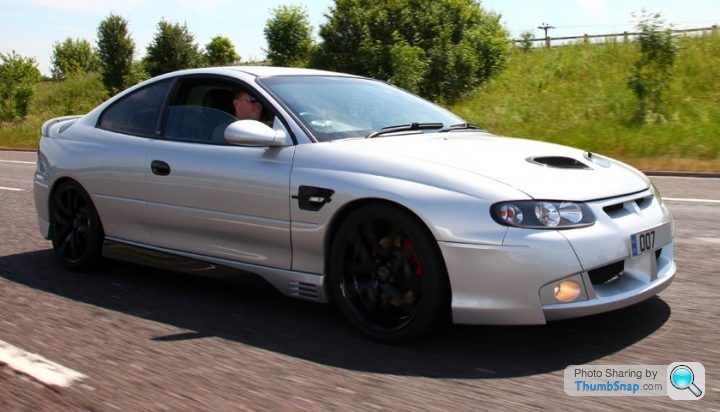 Monaro Vxr Pistonheads - The image depicts a scene of a silver sports car driving down a road. The car is white with black accents, particularly noticeable around the wheels and at the front and rear. The interior of the car is visible, showing a man seated and driving, a side view mirror indicating this. The road on which the car is driving is wide and tarmacked, with several trees visible on the right side of the image. There are no other vehicles in sight.