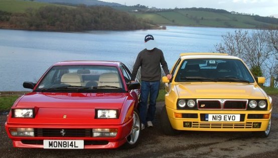 Places to take good photos... - Page 1 - South Wales - PistonHeads - The image depicts a serene setting by a lake with two convertible cars parked on the grass. A person is standing between these vibrant, brightly colored vehicles. The radiant red car has a personal license plate with the letters "MON81L," while the bright yellow car's license plate reads "NI9 EVO." The person is dressed in a sports cap and a dark top, and they appear to be posing for the shot. The background consists of the vast expanse of the lake and rolling hills dotted with trees, adding to the tranquil scene.