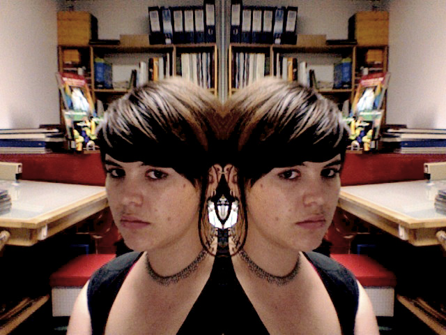 The image features a person looking directly at the camera. The person is seen from the side and in mirror reflection, creating a somewhat symmetric effect. The person has short hair and appears to be somewhat melancholic with a slight frown. They are wearing a sleeveless top and the background is filled with books, suggesting a study or library environment.