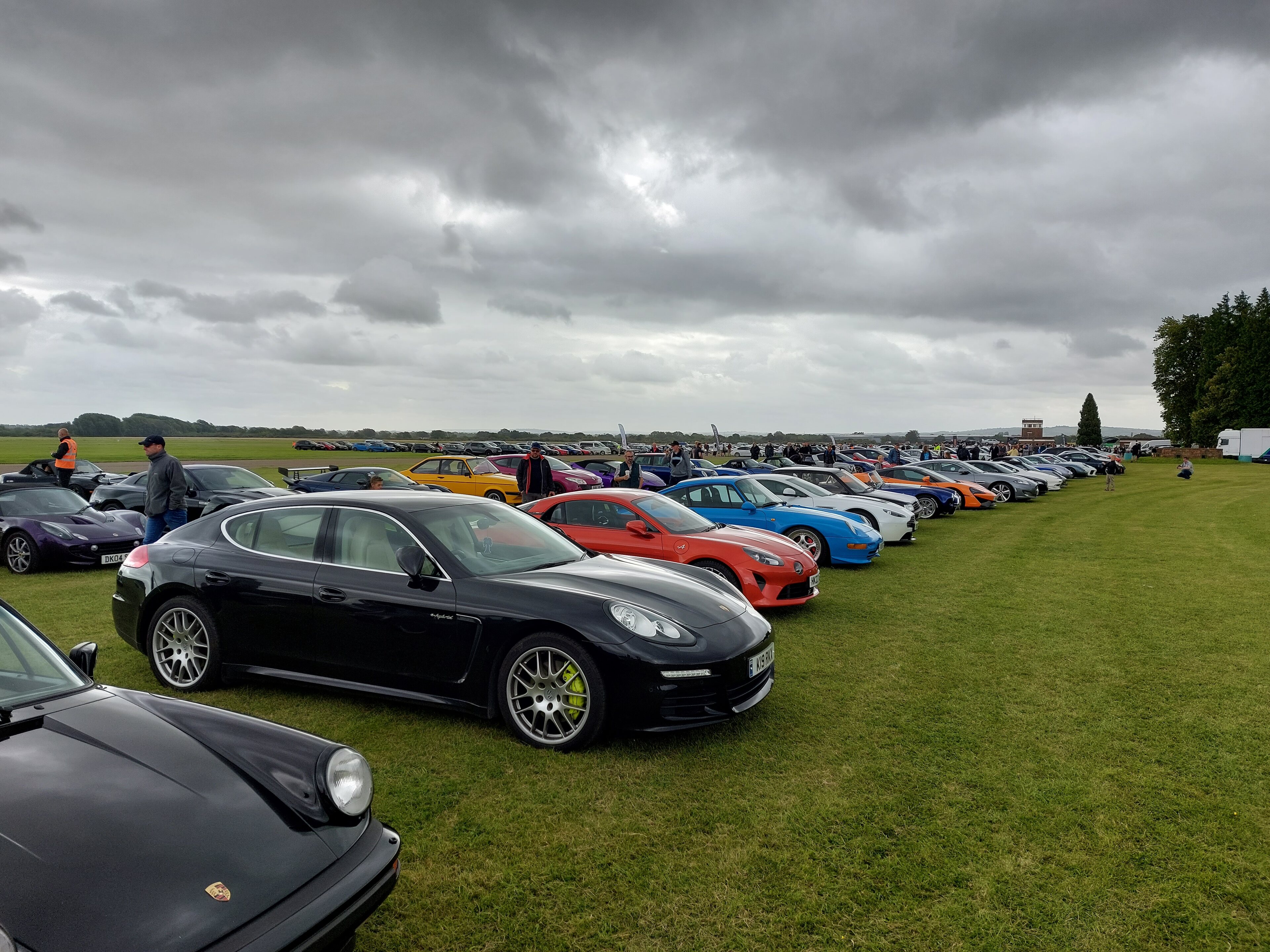 The image captures an outdoor scene, where a large field serves as a parking lot for various cars. The field is filled with rows of parked cars, showcasing a diverse collection of models and colors. The cars are parked in neat lines, indicating a well-organized event or gathering. In the foreground, there's a paved area that provides a contrasting background to the grassy field. The sky above appears overcast, suggesting it could be an overcast day.