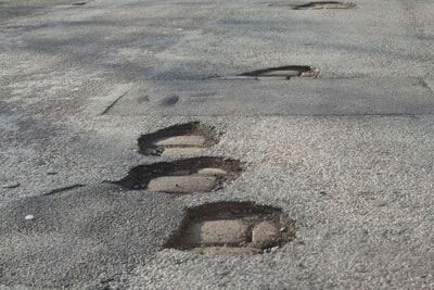 Appalling Pistonheads Roads State - The image captures an urban scene where a grey asphalt road has experienced significant damage. There are four large potholes in the road, each having spread to become smaller holes nearby. Interestingly, the ground appears to be levelling out, suggesting that the road could be undergoing repairs or maintenance. The overall scene conveys a sense of dilapidation and the need for road repair.