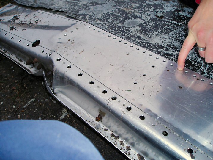 Hunting Pistonheads Plane Wreck Crash - The image shows a close-up of the bottom side of a large flat metal surface, possibly a roof or a plank. The metal has a texture of screw heads, suggesting it is designed for fastening purposes. The surface appears weathered and worn, hinting at exposure to outdoor elements. A person's hand can be seen pointing to one of these screw heads, indicating the focus of the image.