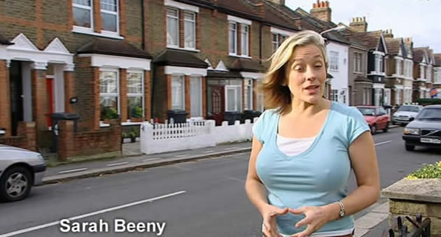 Pistonheads - The image is a television style screenshot featuring a woman standing in a residential street surrounded by cars and a row of houses. The woman is in the foreground, dressed in a light blue top, her hands clasped together, and facing the viewer directly. She has fair skin and her hair is blonde and flows in the wind. The environment suggests a quiet suburban area with houses of similar design. There's a watermark or text at the top that reads "Sarah Beeny."