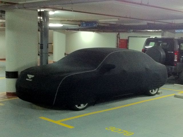 Local customs . . . - Page 1 - Middle East - PistonHeads - The image features a parking garage with cars partially covered by large black car covers. In the foreground, a car is driving towards the viewer while a larger, dark car is positioned further back but nearly filling the frame. The car covers are partially illuminated by lights above, highlighting their presence. The perspective of the photo suggests it may have been taken on a nearby street, as the side angles slightly away from the car covers. The car covers appear to be removed or folded back on the car in the foreground, revealing the car underneath.