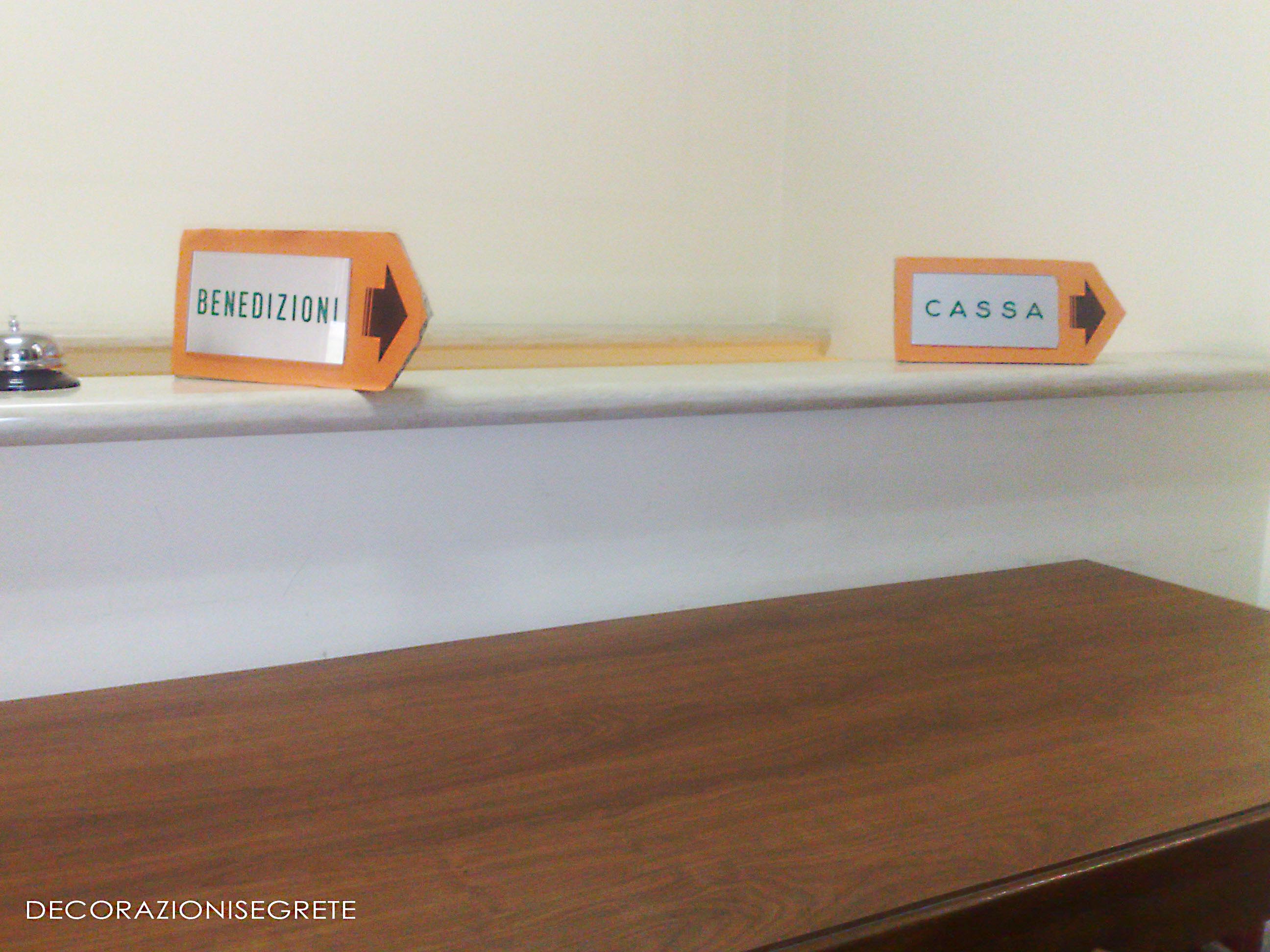 The image depicts a simple, wooden whiteboard placed against a wall. On the left side, there is a small, box-like sign with a green arrow pointing towards a bell. The sign has Italian text and translates to "Call the bell to summon help." On the right, another sign is also small but rectangular, with an orange arrow symbolizing directing to "Cassa." The whiteboard is positioned in a corner, and a table is adjacent to it, suggesting a communal area for announcements or information. The text "DECORAZIONESEGRETE" in Italian at the bottom indicates that the image may have been taken in Italy.