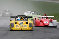 Pistonheads Bikesports - The image captures an intense moment from a track race featuring vintage cars. Two distinct cars, one yellow and the other red, are driving down the road, jockeying for what appears to be the lead. They are the only two in the foreground, though blurred figures of other cars can be seen in the background, adding to the sense of competition. The license plates on the cars are clearly visible, adding a personal touch to the professional setting of the race.