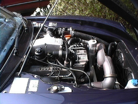 Chimaera Tvr Start Pistonheads Hot - The image depicts the underside of a dark-colored car, likely a sports car given the visible hood vents and the angular design. The engine is exposed through an open bonnet, revealing a complex arrangement of metal pipes and hoses. On the right side of the image, there is a rectangle with the word "Here" inside it, perhaps indicating a specific point of interest or location within the engine bay. The background is out of focus, but it appears to be an outdoor setting, possibly a car show or exhibition.