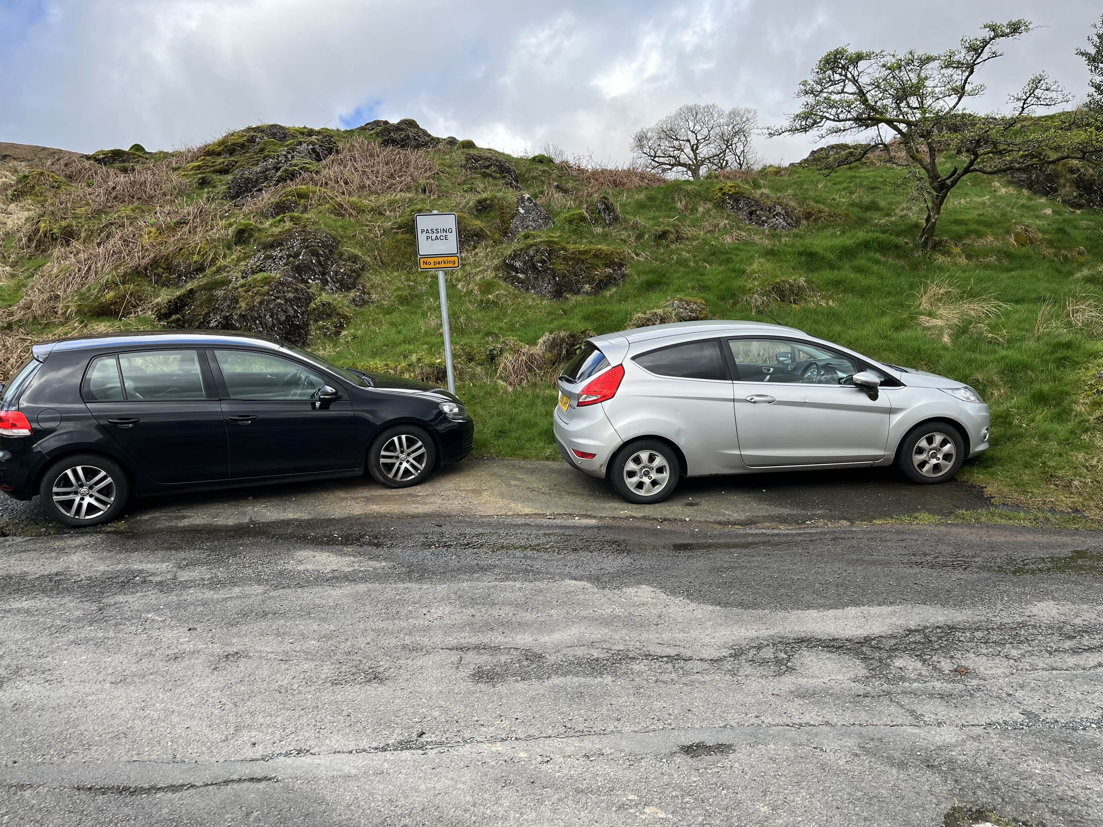 Pistonheads - The image shows a rural setting with two cars parked on the side of a road. The cars are positioned opposite each other, suggesting a parking lot or similar location. There is a grassy hill in the background and a clear sky above. In the foreground, the ground appears to be wet, possibly indicating recent rainfall. A signpost is visible in the background, though the text on it isn't legible. The overall scene conveys a quiet, possibly remote area.