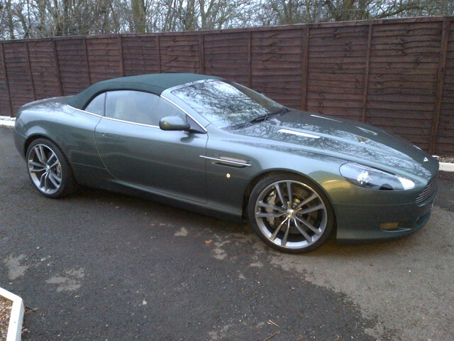 DBS wheels on DB9 - (more or less) - Page 1 - Aston Martin - PistonHeads - The image features a gray convertible sports car parked in front of a brown wooden fence. The car is sleek and modern, with a tilted roof and a single, thin blade of air resistance over the sleek body. It stands out against the dark red street it's parked on, which is lined with a small white chalk border. The fence provides a contrasting background to the car's reflective surface, and the overall scene suggests a quiet, suburban setting.