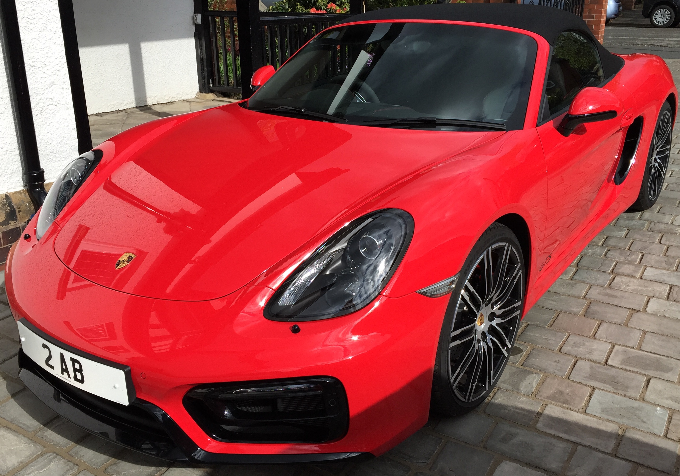 A red and black car with a red umbrella - Pistonheads - The image depicts a vibrant red Porsche sports car parked on a tiled driveway. The car is positioned at an angle, allowing a clear view of its sleek design. The Porsche's curved body and distinct trapezoidal logo are instantly recognizable, suggesting a high level of craftsmanship and performance. The black door handles, side mirrors, and roof add a contrast to the bright red paint, enhancing its aggressive and powerful aesthetics. The Porsche's license plate reads "2 AB", adding a personal touch to the vehicle. The stone driveway provides a suitable backdrop, highlighting the car's vibrant color and sleek design.