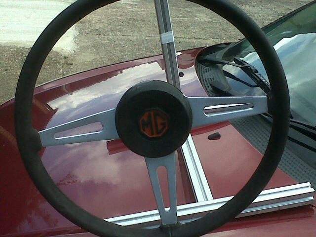 14" or 15" steering wheel mgb - Page 1 - MG - PistonHeads - The image shows the interior of a red car, focusing on the steering wheel. The steering wheel appears to be a three-point format, commonly found in many vehicles. On the wheel, the emblem of the General Motors Company is visible, indicating the manufacturer of the car. The seat belt is not attached to the wheel, and the wheel has a black cover. The car's interior is not in full view, but the red color of the car is contrasted by the gray exterior.