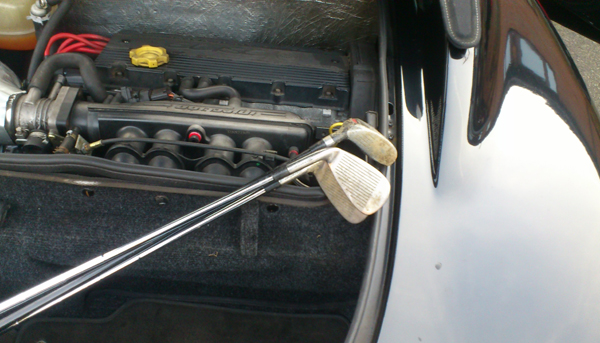 Pics of your bootspace pls - Page 1 - Elise/Exige/Europa/340R - PistonHeads - The image shows a close-up view of a golf club resting near the engine of a car. The car has a shiny surface which reflects light, and the engine components, such as tubes and hoses, are visible. The golf club is positioned at an angle, and the brand and model of the club are not discernible. The setting suggests a parking area or an outdoor storage location. The image captures a contrast between the man-made technology represented by the car and the leisure activity symbolized by the golf club.