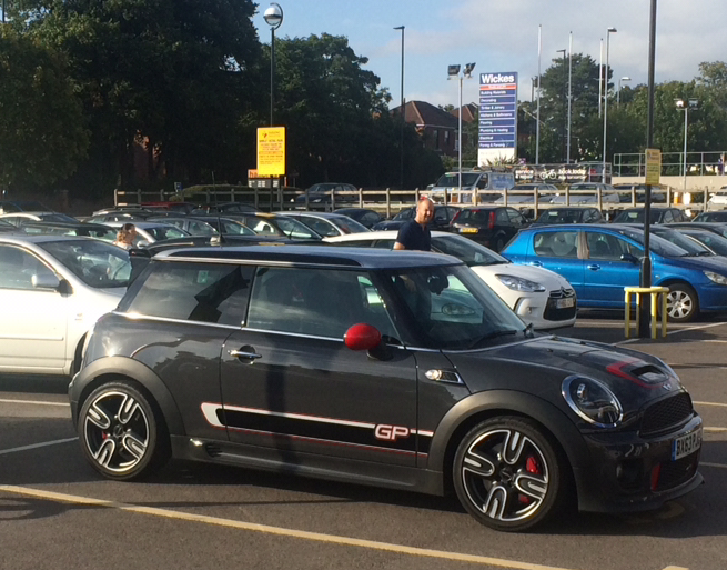 Stickering up for 2016 - Page 1 - Le Mans - PistonHeads - The image depicts a parking lot scene. Dominating the frame is a black Mini Cooper. It's parked angled to the right of the lot, with other cars visible in the background. A noticeable feature of the car is a red aluminum ball on the left side. A person is standing near another car, perhaps engaged in conversation or supervision. The environment is structured with lampposts and signs, typical of a public parking area. The setting suggests it could be a sunny day given the shadows and the overall lighting.